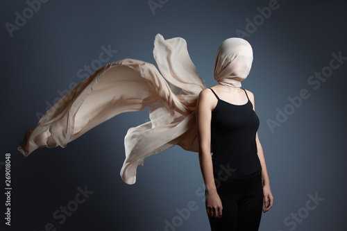 An anonymous model, is covered by a beige veil. A woman in a black top with her head wrapped in a fluttering veil on a dark gray background