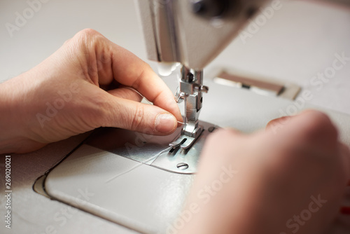 Woman's fingers putting thread in needle loop on electric modern sewing machine for start sewing. Steel needle with looper and presser foot. Close up view of female working hands. Blurred background.