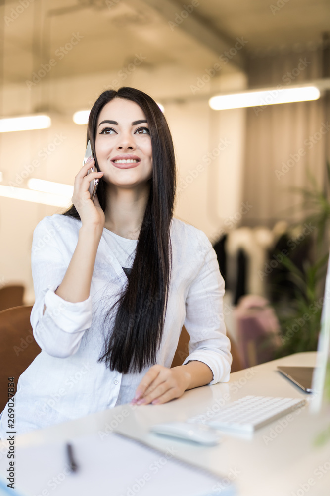 Portrait of a smiling businesswoman sitting at a white desk talking on the phone touching some corporate papers her laptop next to her Portrait of a smiling businesswoman sitting at a white desk talking on the phone touching some corporate papers her laptop next to her