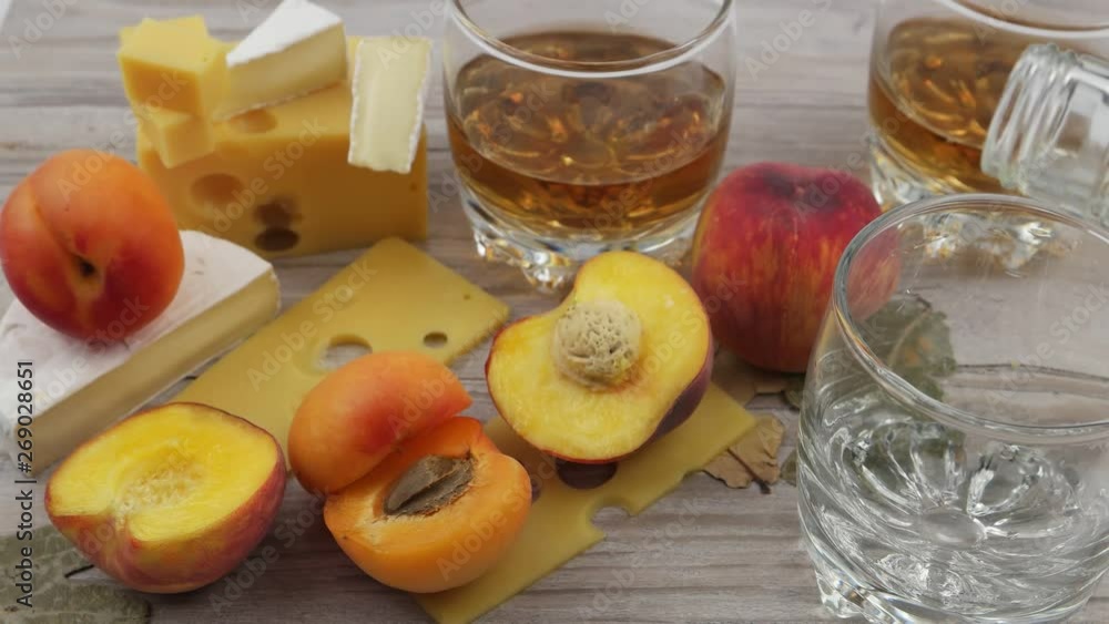 A glassful is filling with whiskey. Snack to complement to whiskey party. Glasses of whiskey, pieces of swiss cheese and cheese brie, fruits and leaves of oak on a wooden plank. A glassful is filling with whiskey. Snack to complement to whiskey party. Glasses of whiskey, pieces of swiss cheese and cheese brie, fruits and leaves of oak on a wooden plank.