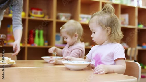 little kids are sitting at the table, they eat something from the plate by themselves using spoon, after that someone brings them drinks in mugs and puts them on the table, in the background is a