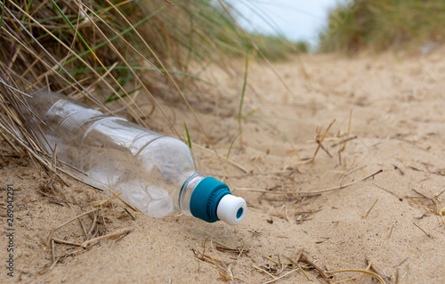 Discarded plastic drinks bottle lies amongst sand and sedge grass in dunes at a beach