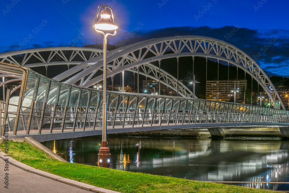 Obraz premium puente metálico blanco iluminado de noche con reflejo de las luces en el agua del rio con una farola en la vista