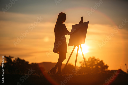 Silhouette of a blonde girl paints a painting on the canvas with the help of paints. A wooden easel keeps the picture. Summer is a sunny day, sunset © Aleksandr