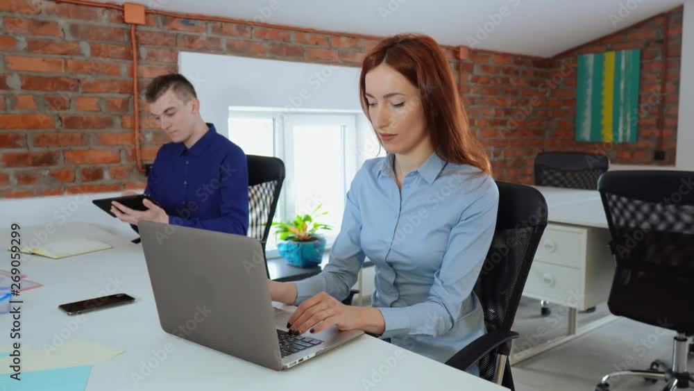 young caucasian coworkers sitting at the workplace in loft office. man using digital tablet reading documents woman with red hair typing on laptop looking in the screen computer then to the camera young caucasian coworkers sitting at the workplace in loft office. man using digital tablet reading documents woman with red hair typing on laptop looking in the screen computer then to the camera