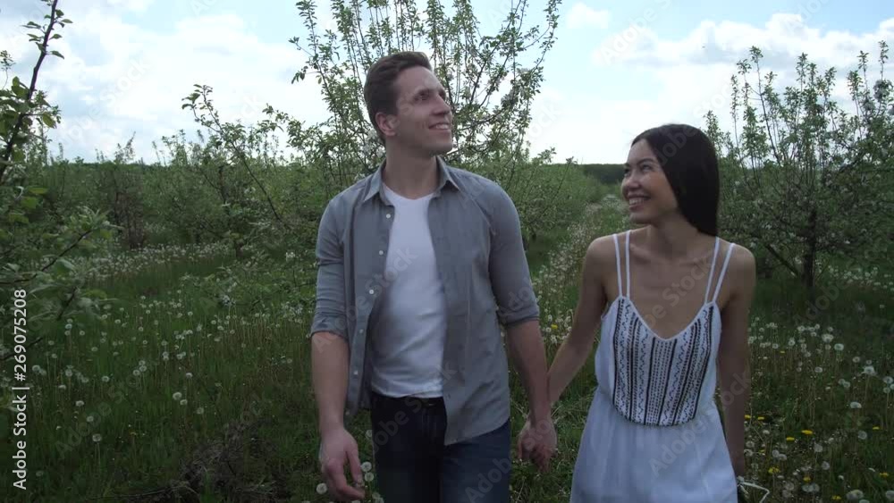 Close-up of beautiful mixed race couple dating in spring blooming orchard, man embracing beloved woman holding bouquet of flowers. Happy young bride and groom strolling among flowering apple trees Close-up of beautiful mixed race couple dating in spring blooming orchard, man embracing beloved woman holding bouquet of flowers. Happy young bride and groom strolling among flowering apple trees