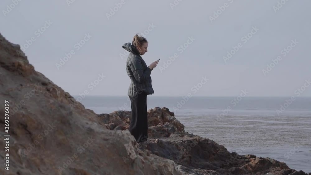 long shot of girl standing and looking into her smartphone on the rocky shore long shot of girl standing and looking into her smartphone on the rocky shore