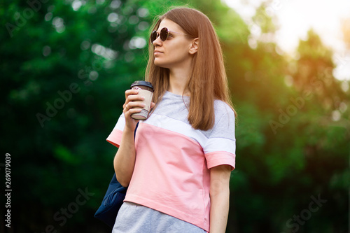 Pretty student girl in sunglasses drinking coffee at summer park
