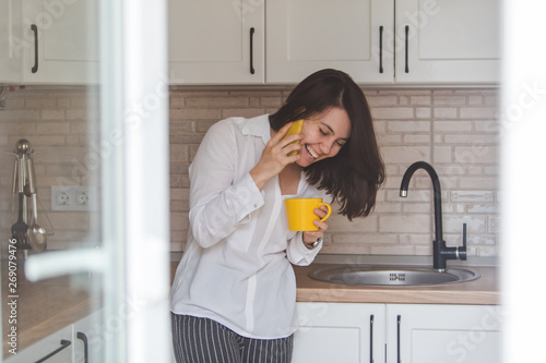 smiling happy woman talking no phone drinking tea from yellow mug at the kitchen