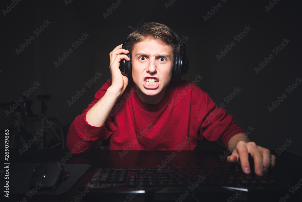 Emotional young man playing video games on a computer at home, wicked looking computer screen.Aggressive guy in the headphones plays a video game on a computer with an evil eye looking at the monitor