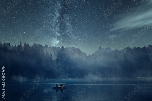 Nachtangeln auf einem See bei Nacht mit Sternenhimmel und Nebel. Im Hintergrund Wald, Berge und die Milchstraße