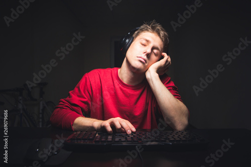 Sleepy guy in headphones and in a red sweatshirt sits at home at a computer and is tired of sleep. Gamer fell asleep from a boring game. Teenager uses a computer and sleeps