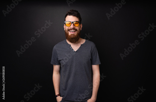 Photo of bearded guy wearing stylish sunglasses, smiling at camera over dark background