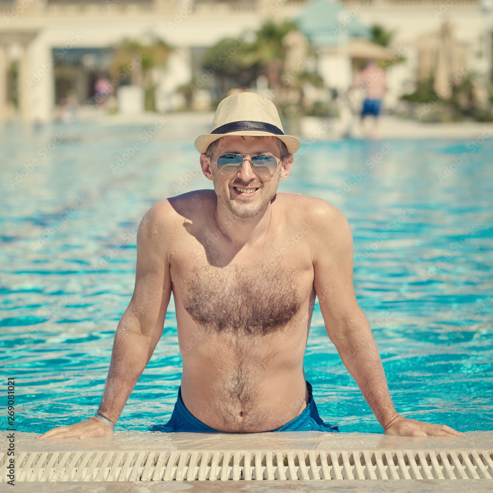 Happy man in sun hat and sunglasses on side of swimming pool on summer vacation.