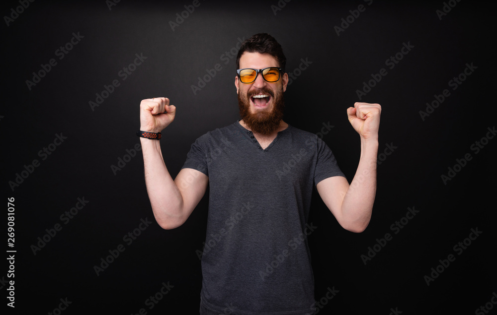Naklejka premium Photo of bearded man wearing sunglasses celebrating with rised hands standing over dark background