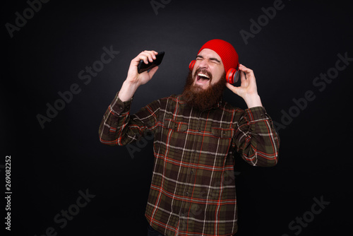 Photo of happy bearded guy listening music on headphones, standing over dar isolated background © Vulp