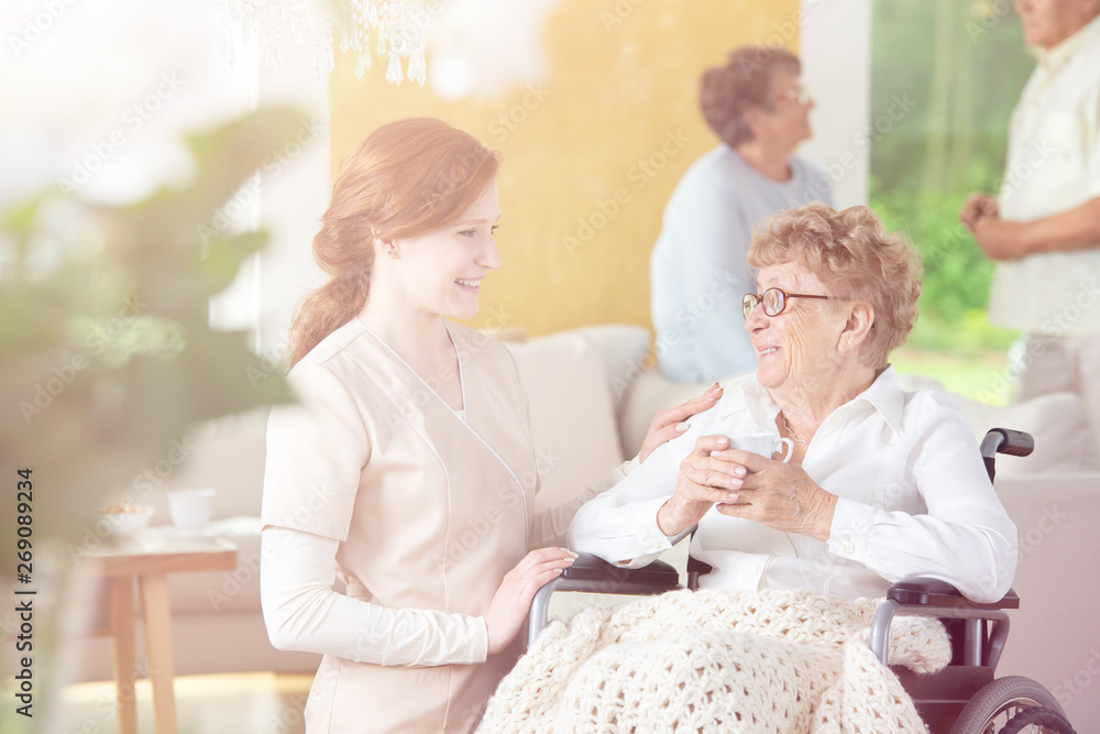 Friendly caregiver helping smiling senior woman in the wheelchair in the nursing house Friendly caregiver helping smiling senior woman in the wheelchair in the nursing house