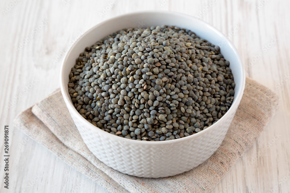 Dry green french lentils in a bowl over white wooden background, side view. Dry green french lentils in a bowl over white wooden background, side view.