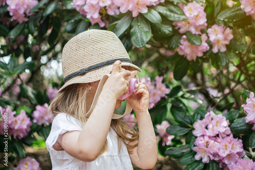 Cute little girl in white sunglasses playing with children's wooden camera in the park with rhododendron flowers