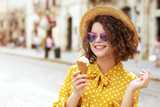 Outdoor close up portrait of young happy smiling cute woman wearing straw hat, colorful heat sunglasses, polka dot dress, enjoying an ice-cream, posing in street of European city. Copy, empty space
