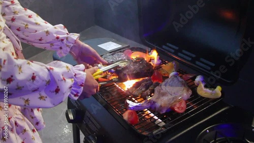 Sexy young girl prepares steaks and grilled vegetables on the roof of the house overlooking the night city, the wind lifts her dress and underwear can be seen