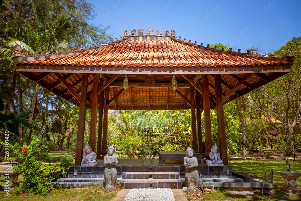 Antique gazebo pavilion with a roof asian style pagoda. In a summer tropical garden. A stone path along which the statues stand leads to the building. Inside the area of rest and meditation.