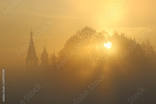 Rising sun rays shining through trees folage and mist with background of church silhouette and yellow morning sky