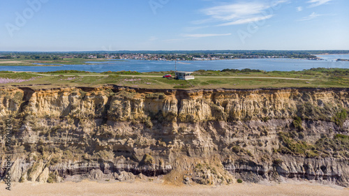 An aerial view of a coastguard tower on the top of a hill with eroded cliff along a sandy beach with an harbor in the background under a majestic blue sky and some white clouds