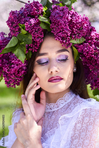 Portrait of a young beautiful girl in a wreath of lilacs. White lace dress on the perfect figure of the model. Photo session in the spring garden in the open.