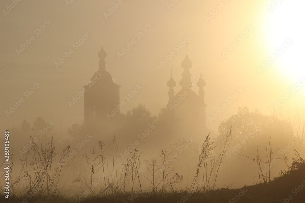 Misty rural landscape in rising sun light with silhouettes of church and grass 