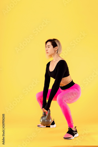 Fitness woman in fashionable pink and black sportswear work out with kettlebell on yellow background. Strength and motivation.