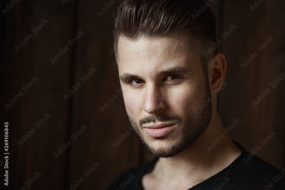 Muscular model young man with beard in black t-shirt on dark wooden background. Fashion portrait of brutal sporty sexy strong muscle guy with modern trendy hairstyle. Model, fashion concept. Muscular model young man with beard in black t-shirt on dark wooden background. Fashion portrait of brutal sporty sexy strong muscle guy with modern trendy hairstyle. Model, fashion concept.
