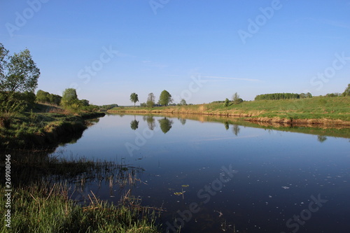 Wide calm river with green shores and rare trees reflects blue summer sky