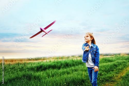 Little girl, launches a toy plane, into the air against, the background of green grass. Child launches a toy plane. Beautiful little girl, stands on the grass and launches a pink toy plane.