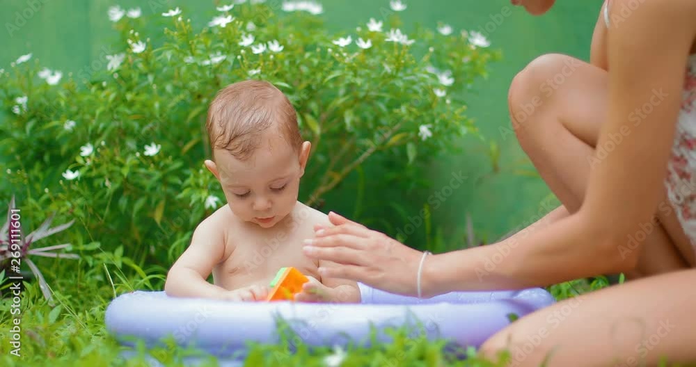 A little boy of 8 months old is bathed in a mini pool in the courtyard on the green grass. The baby is splashing and laughing with joy. Mother sits next to the baby and plays with him. real time