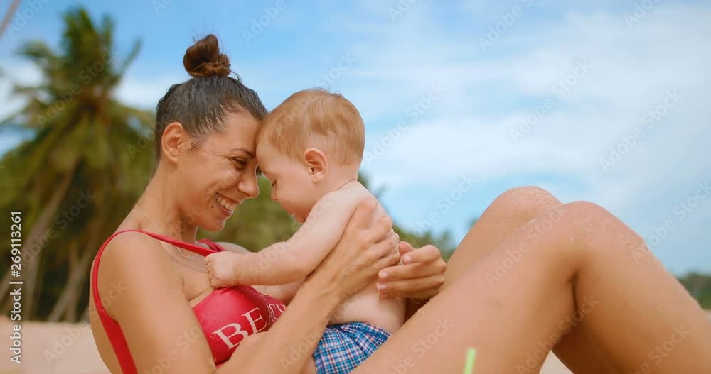 Mom with a baby sitting on the sand on the beach hugs and kisses the child. in slow motion Mom with a baby sitting on the sand on the beach hugs and kisses the child. in slow motion