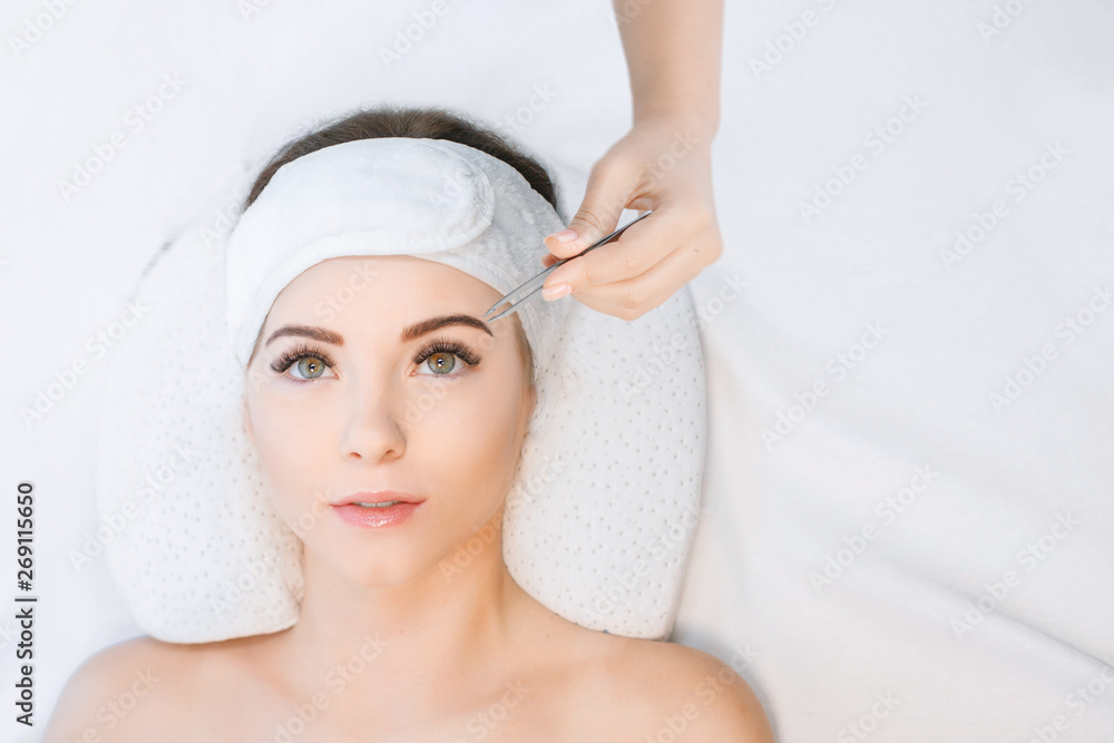 Beautiful young girl with long eyelashes tweezing her eyebrows in a beauty salon. Woman doing eyebrow permanent makeup correction . Microblading brow.