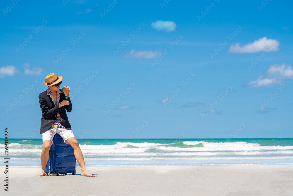 Wide shot selective focus of young businessman traveler in casual wear sitting on blue suitcase on the island beach in summer day holidays vacation with blurred blue sea, sky and clouds backgrounds Wide shot selective focus of young businessman traveler in casual wear sitting on blue suitcase on the island beach in summer day holidays vacation with blurred blue sea, sky and clouds backgrounds
