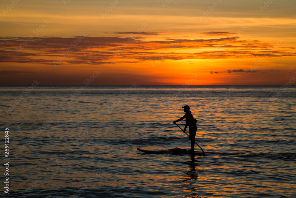 Silhouette of young man paddling on a SUP board in the sea at sunset, rear view