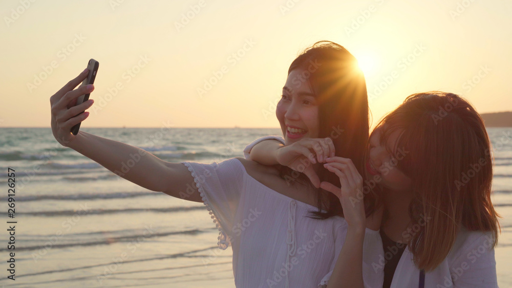 Young Asian lesbian couple using smartphone taking selfie near beach. Beautiful women lgbt couple happy relax enjoy love moment when sunset in evening. Lifestyle lesbian couple travel on beach concept