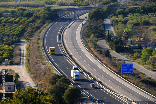 A long curve of the AP7 motorway in Spain near Denia, photographed from above with cars and trucks. On the right is a blue sign for the next departure.