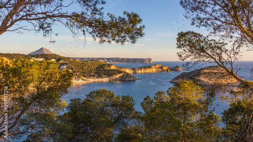 A wonderful view over the cliffs and bays of the northern Costa Blanca near the city of Javea in the sunshine and blue sky with trees and branches in the foreground. photo