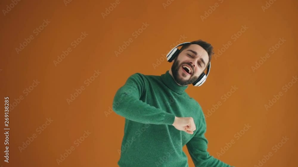 Slow motion portrait of joyful bearded guy dancing and listening to music in wireless headphones enjoying modern technology moving on orange background. Slow motion portrait of joyful bearded guy dancing and listening to music in wireless headphones enjoying modern technology moving on orange background.