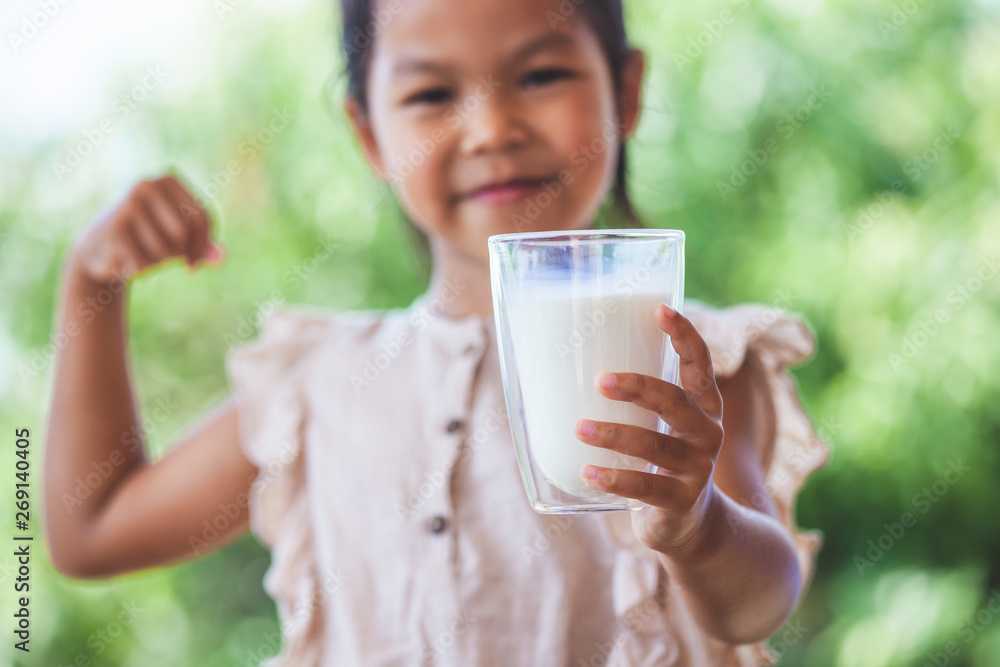 Cute asian child girl holding glass of milk and make strong gesture on green nature background Cute asian child girl holding glass of milk and make strong gesture on green nature background