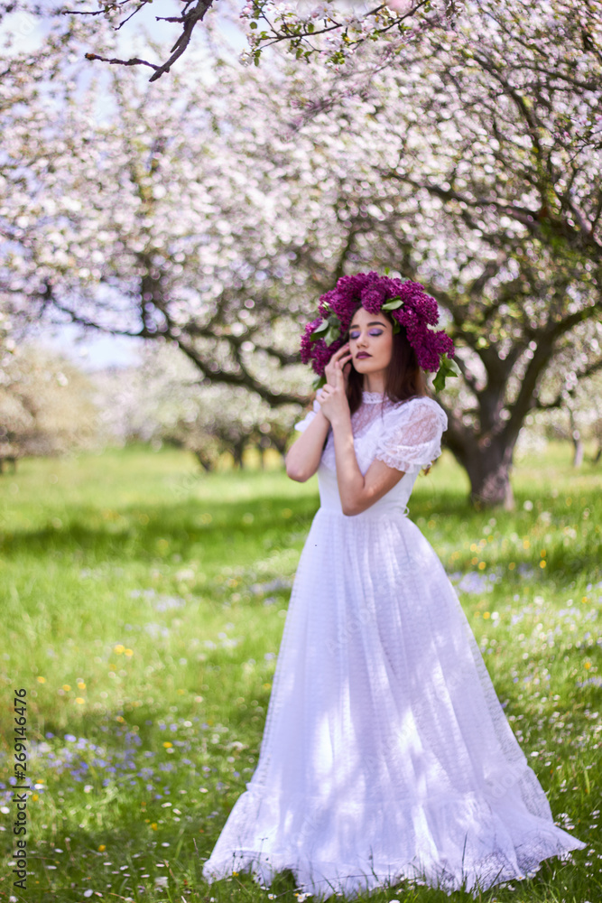 Young beautiful girl in a wreath of lilacs. White lace dress on the perfect figure of the model. Photo session in the spring apple orchard in the open.