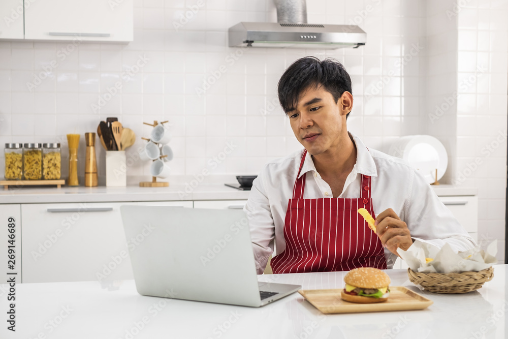 Asian man in white shirt and red apron in a white kitchen holding french fried and using a laptop
