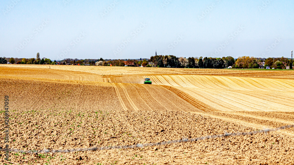 Agricultural farmland with a tractor plowing land with trees and villages in background against blue sky, preparing land to grow potatoes, sunny day in Oensel South Limburg, Netherlands Agricultural farmland with a tractor plowing land with trees and villages in background against blue sky, preparing land to grow potatoes, sunny day in Oensel South Limburg, Netherlands