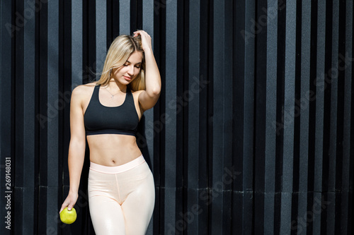A young, sympathetic blonde woman is actively spending time outdoors, holding a green apple. The girl is dressed in sportswear. Time for a healthy snack. Health and benefits for the body