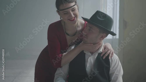 Handsome confident mafia boss in a hat and a vest sitting in an abandoned building. The elegant woman in red dress coming from behind and hugging the man smiling. Happy couple of gangsters photo