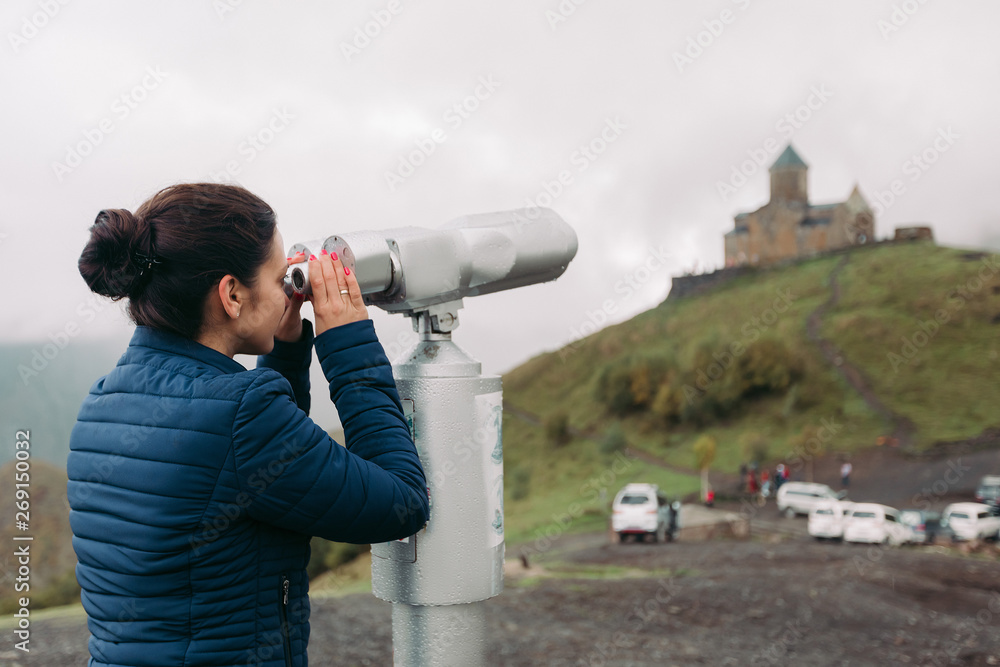 Young tourist woman looking through coin operated high powered binoculars on the Caucasus Mountains, Kazbegi, Georgia